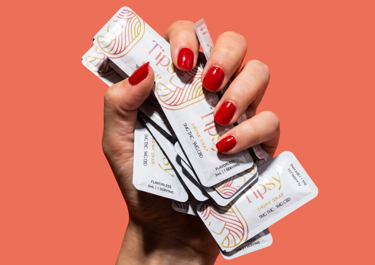 A hand with red manicured nails holds several Tipsy Drink Snaps packets against a solid coral background.
